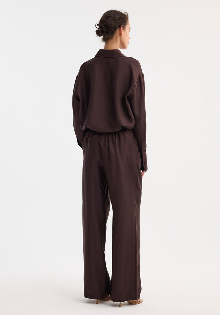 Woman wearing Wide leg silk trousers | mahogany, featuring an elastic waistband and drawstring, styled with a tucked blouse and light-tan heeled sandals.