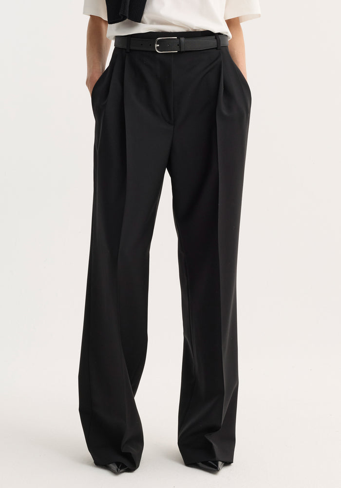 Model wearing mid-waist tailored trousers in black, featuring double pleats, wide legs, and slim belt, crafted from virgin wool-mohair blend for structure and breathability.