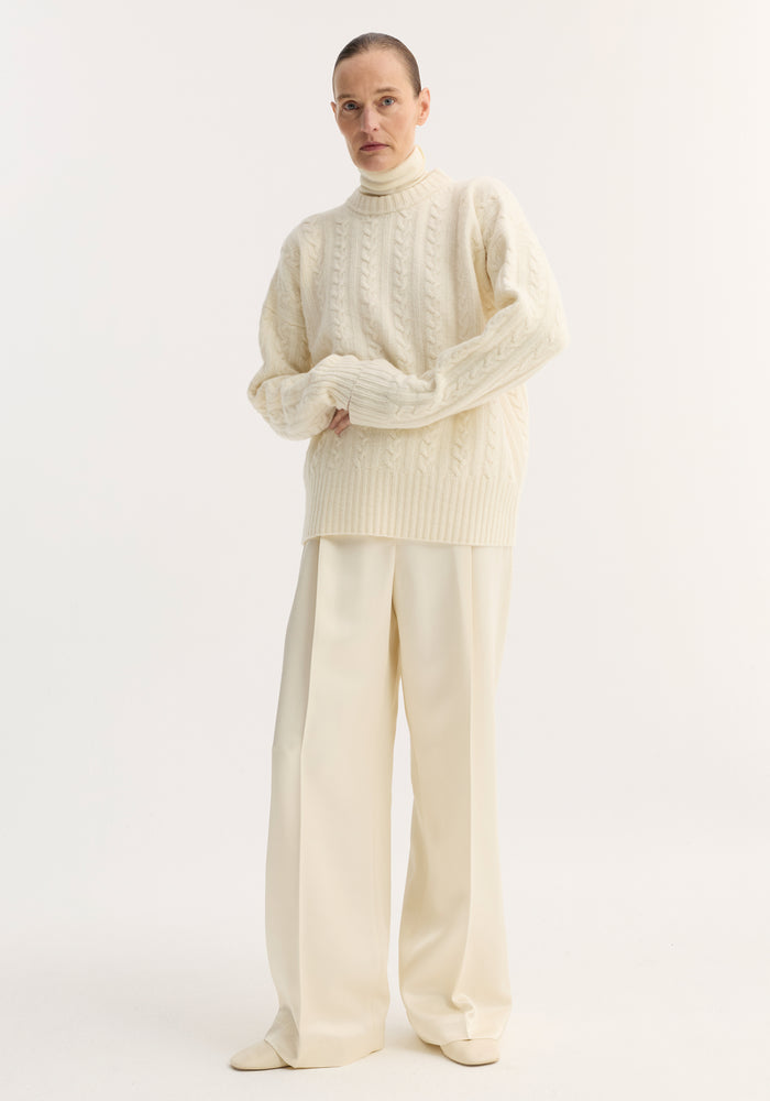 Model wearing Relaxed drawstring satin trousers in cream, featuring a long wide-leg silhouette, drawstring waistband, and back welt pockets, paired with a cable-knit sweater.
