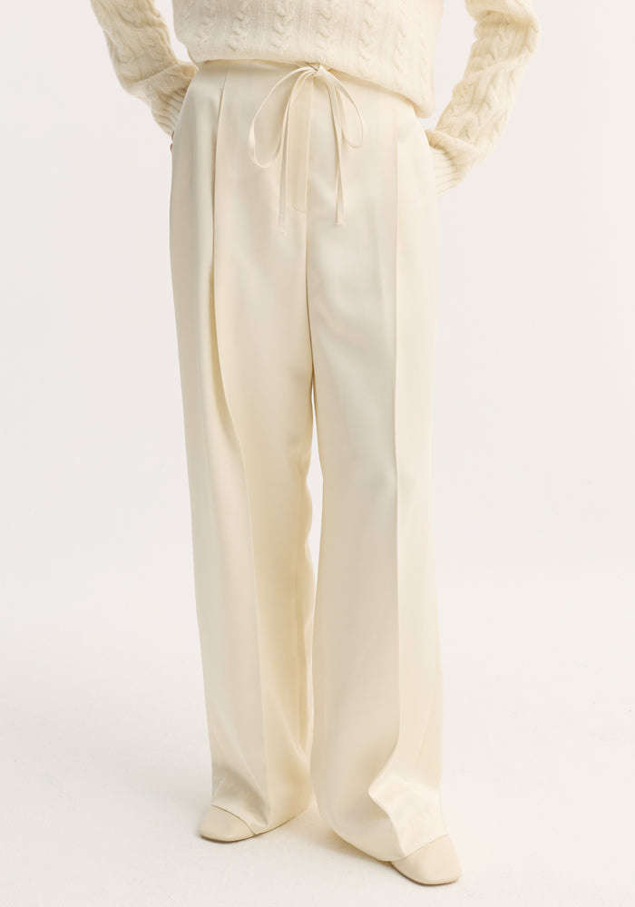 Relaxed drawstring satin trousers in cream, featuring a wide-leg, long silhouette with front pleats and a drawstring waistband, worn with matching shoes.