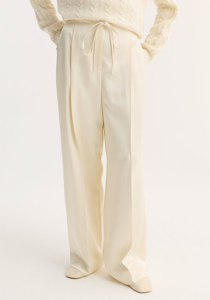 Relaxed drawstring satin trousers in cream, featuring a wide-leg, long silhouette with front pleats and a drawstring waistband, worn with matching shoes.