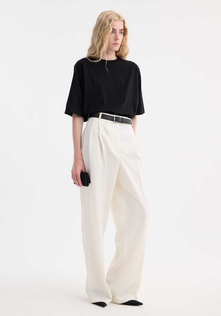Model wearing Mid-waist tailored trousers in soft off white, featuring a wide-leg design, double pleats, and a black leather belt against a neutral backdrop.