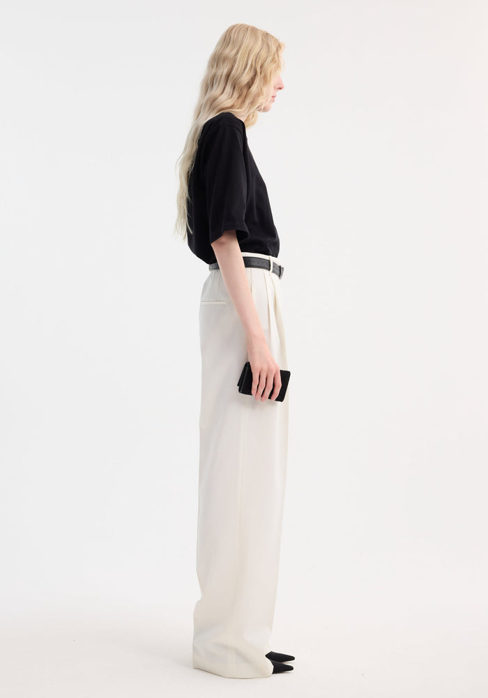 Mid-waist tailored trousers in off white, featuring a high-waist, wide-leg design with double pleats, worn by a tall model holding a small black object.
