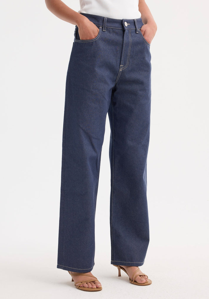 Signature baggy fit raw denim jeans in a high-waist style, straight-leg cut, worn with tan strappy sandals in a studio setting.