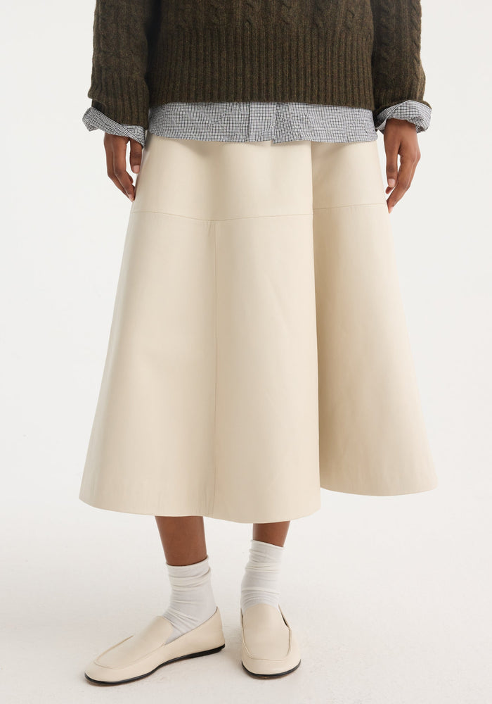 A-line leather skirt in chalk, featuring croco embossing and minimalist detailing, paired with ivory loafers and slouchy socks, highlighting its voluminous silhouette.