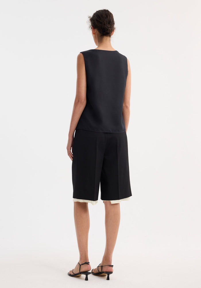 Model wearing Contrast Extended Lining Shorts in black, showcasing tailored waistband, wide legs, and contrasting underlayer. Styled with strappy sandals, hair in a loose bun.