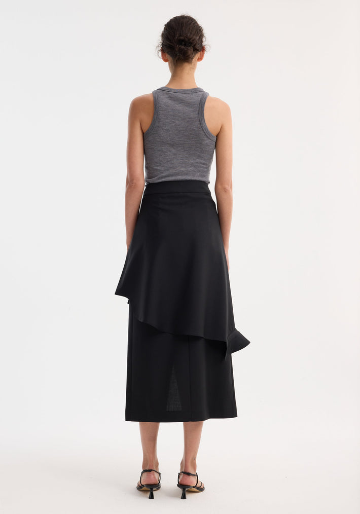 Model in Shirt wrap skirt | black with asymmetrical draped overlay, sleeveless gray tank top, strappy sandals, hair in bun, showcasing skirt's layered design.