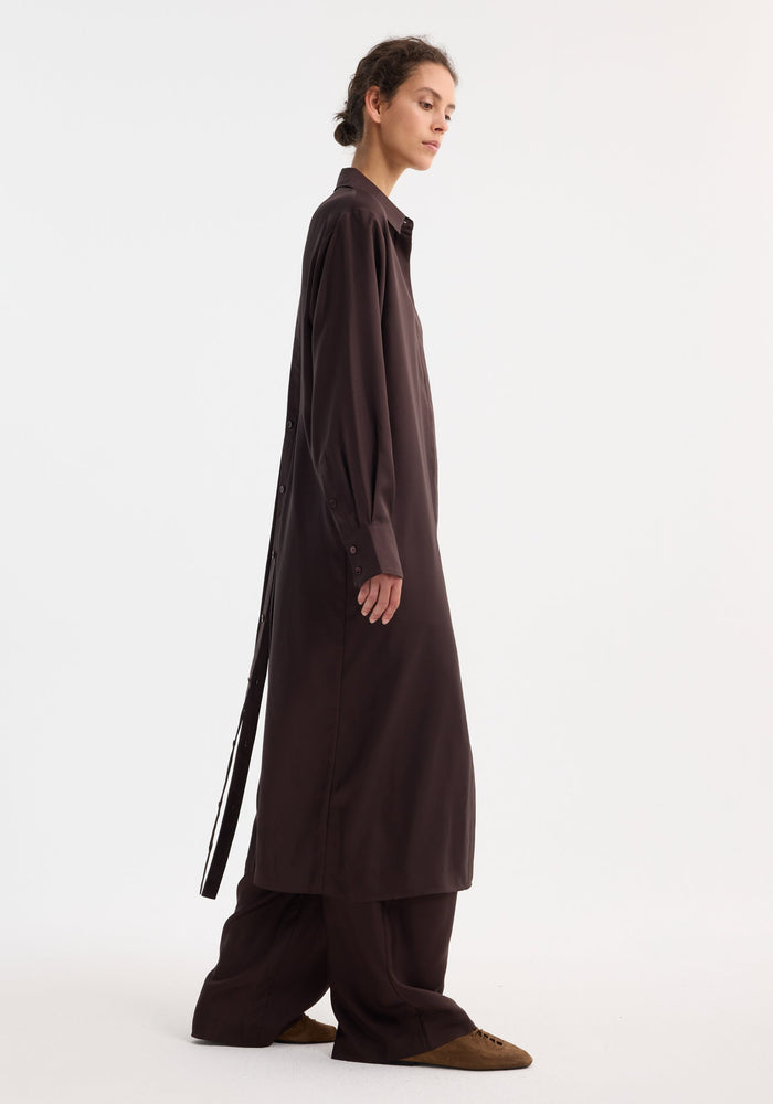 Person in profile wearing a Silk open back wrap dress in mahogany with wide-leg trousers, featuring button closure, wrap tie, and extra long rear hem.