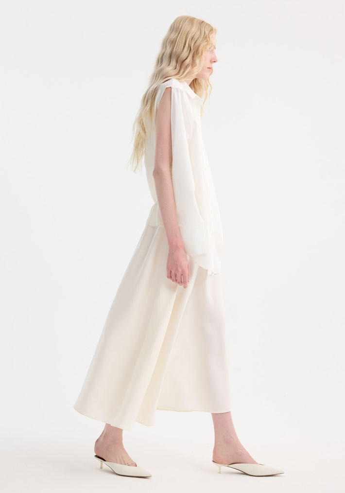Model in Poplin silk combined gilet-dress, featuring a loose sleeveless blouse and flowing skirt, styled with pointed white mules, showcasing a minimalist, monochromatic look.