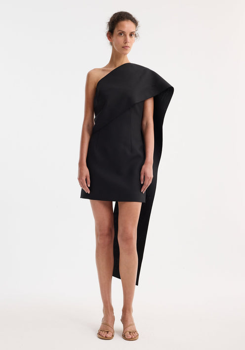 Short sculptural sash dress in black featuring an asymmetric draped shoulder sash, structured bodice, and straight mini skirt, creating a bold, elegant silhouette.