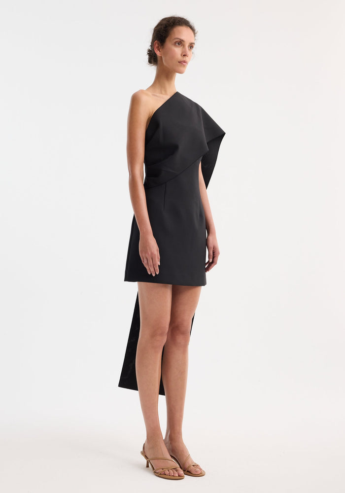Model wearing a Short sculptural sash dress | black, featuring an asymmetrical draped panel and strappy nude sandals, highlighting the dress's structured elegance and minimalist design.