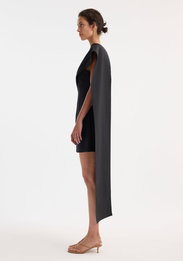 Short sculptural sash dress | black, featuring a side-profile of a woman wearing a fitted black dress with a floor-length cape panel and nude-tan kitten-heel sandals.