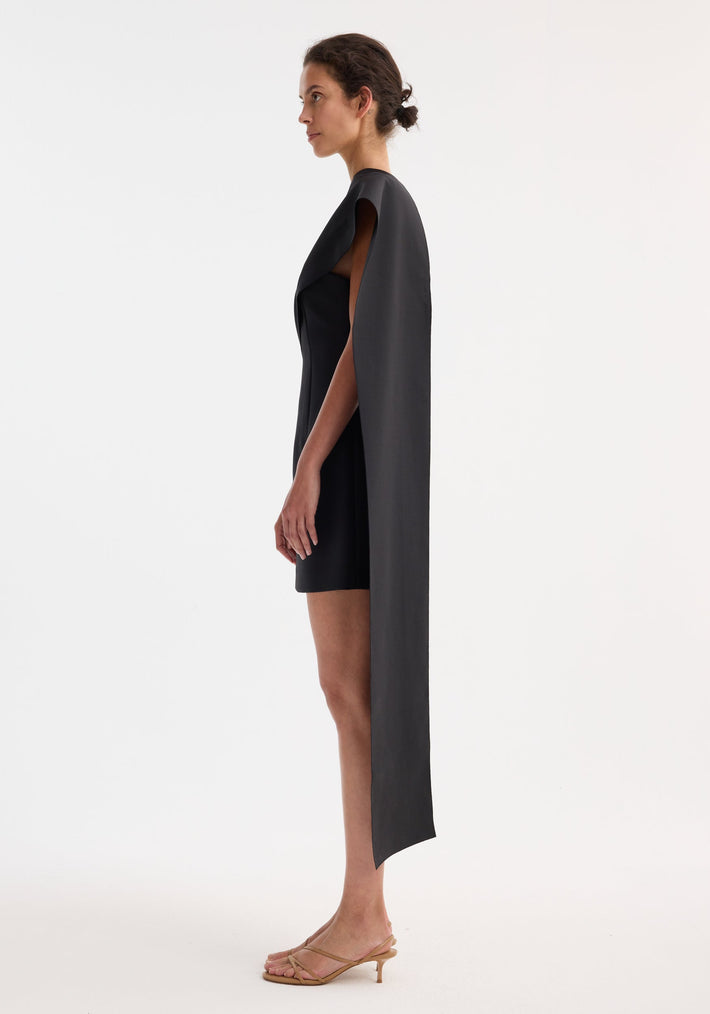 Short sculptural sash dress | black, featuring a side-profile of a woman wearing a fitted black dress with a floor-length cape panel and nude-tan kitten-heel sandals.