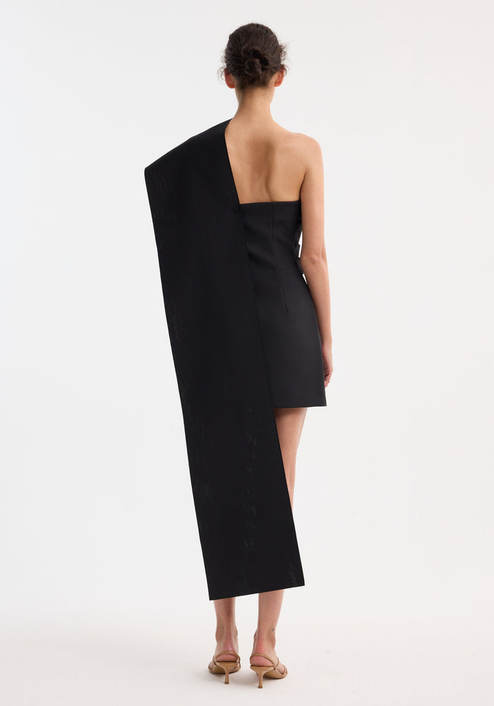 Back view of woman in a Short sculptural sash dress | black, featuring an asymmetrical draped shoulder sash extending to her ankles, paired with nude heels.