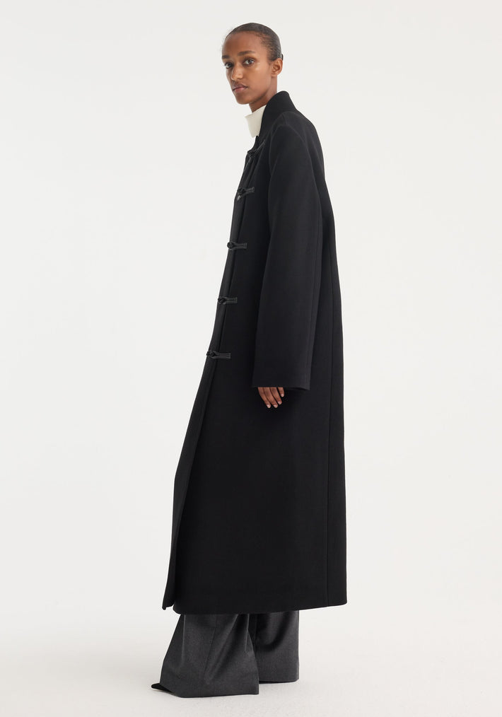 Model wearing a Mandarin wool coat in black, featuring an asymmetrical toggle closure, minimalist long design, and side pockets. Paired with wide-leg gray trousers.