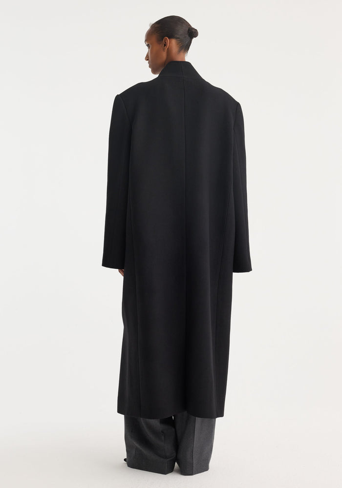 Mandarin wool coat in black, worn long with a high collar. The back view shows a minimalist design, wide-leg gray trousers visible underneath.