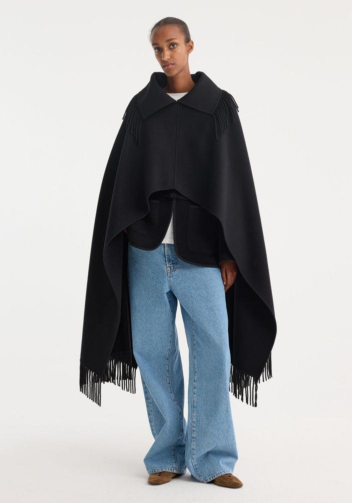 Model wearing Fringe poncho double-faced wool, black, with long fringe details, over a white top and light-wash jeans, paired with brown suede shoes.