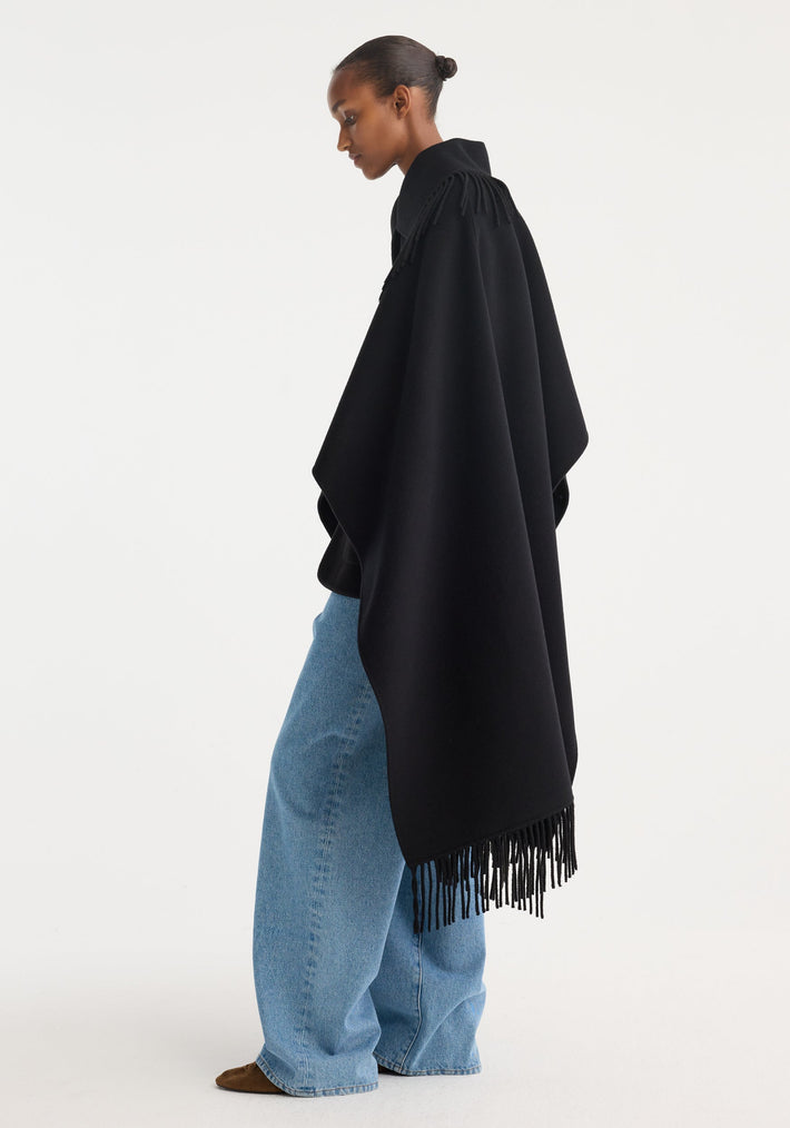 Model in profile wearing a black Fringe Poncho double-faced wool, featuring fringe detailing, over light-blue jeans with brown suede shoes slightly visible.