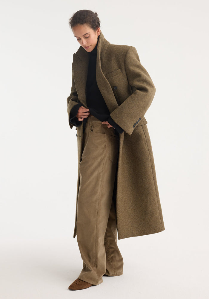 Hourglass wool coat in forest melange on model; features double-breasted design, strong shoulders, feminine waist, styled over a black sweater and tan trousers.