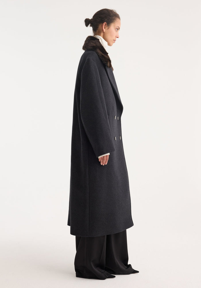 Double-breasted coat with faux fur collar | anthracite melange: Side-profile of a woman modeling a long, tailored wool coat with fur-trimmed collar over a turtleneck and wide-leg trousers.