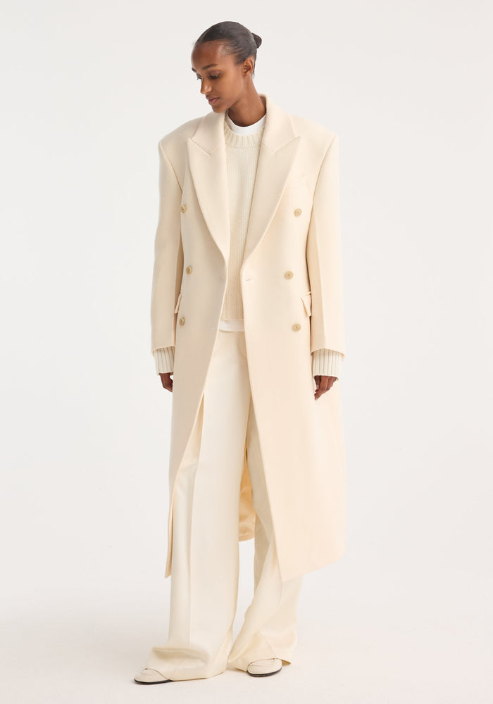 Hourglass wool coat in off white, featuring double-breasted design and wide lapels, worn over a chunky knit sweater and wide-leg trousers, showcasing minimalist elegance.
