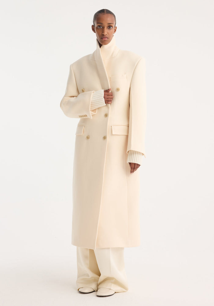 Model wearing Hourglass wool coat, off white, featuring a double-breasted design and strong shoulders, paired with matching trousers and loafers for a minimalist look.