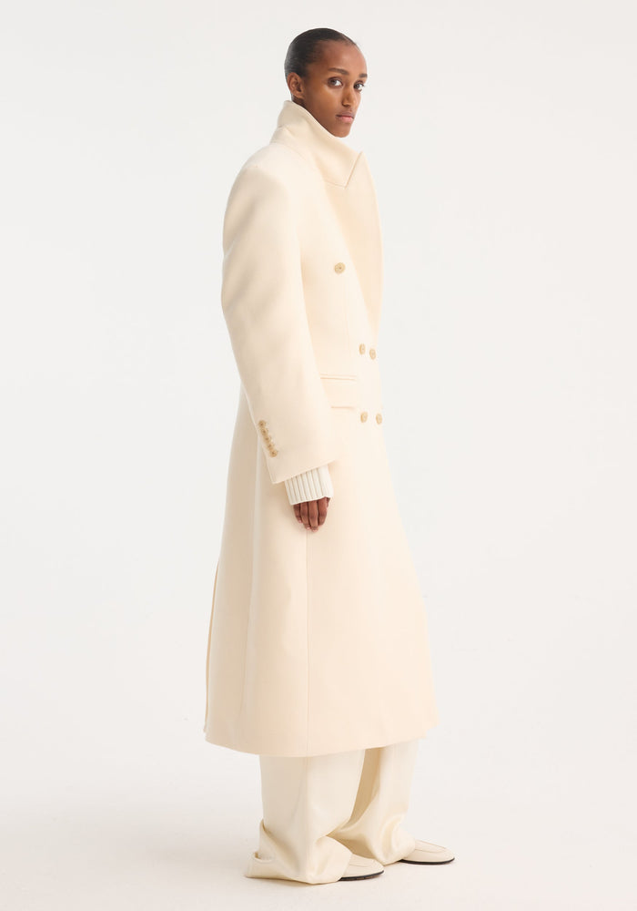 Model wearing Hourglass wool coat | off white, showcasing its long, double-breasted design with strong shoulders and a feminine waist, paired with wide-leg trousers.