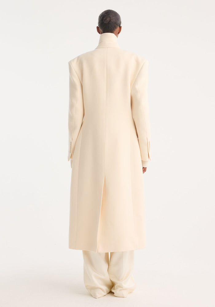 Model in a rear-view wearing an Hourglass wool coat in off white, showcasing a high collar, single back slit, over matching wide-leg trousers.