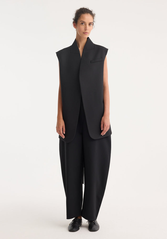 Woman models Reimagined oversized gilet, featuring a sleeveless wool blend front, silk back, wrap-style belt, paired with wide-leg trousers and lace-up shoes.