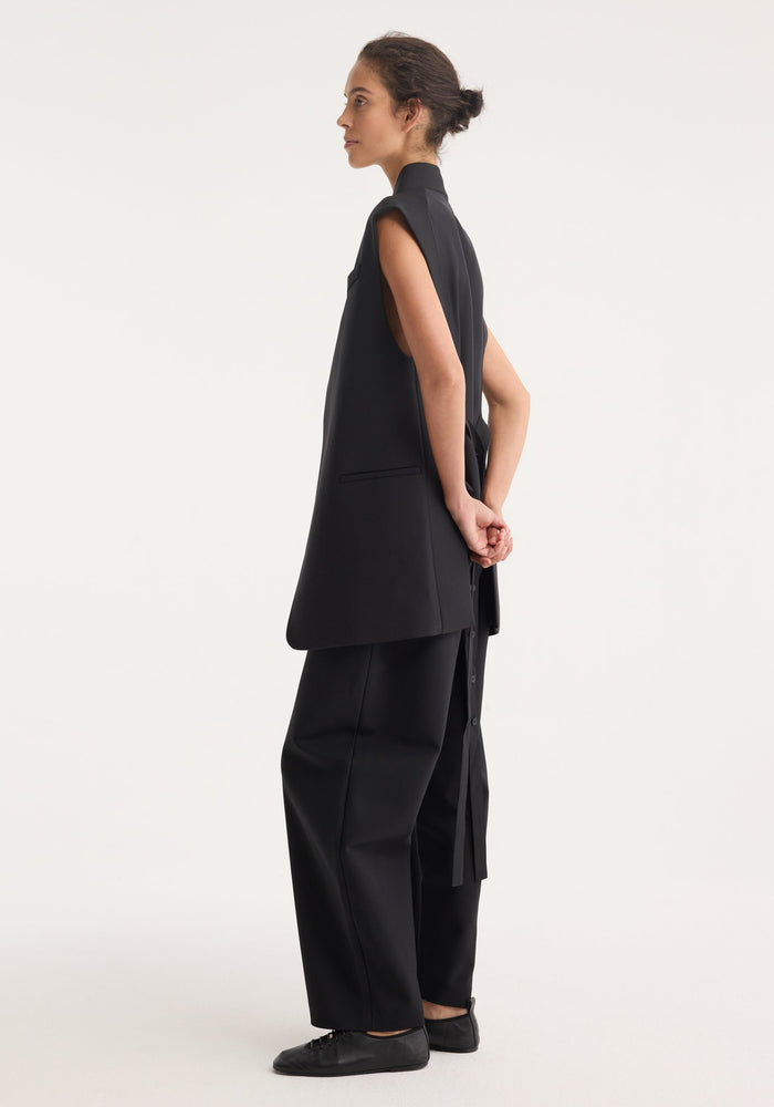 Reimagined oversized gilet in black, featuring a wool blend front, silk back insert, wrap-style belt, worn by model with wide-leg trousers and flats.