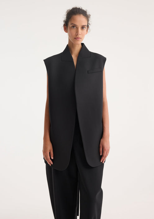 Woman models Reimagined oversized gilet, featuring a sleeveless wool blend front, silk back, wrap-style belt, paired with wide-leg trousers and lace-up shoes.