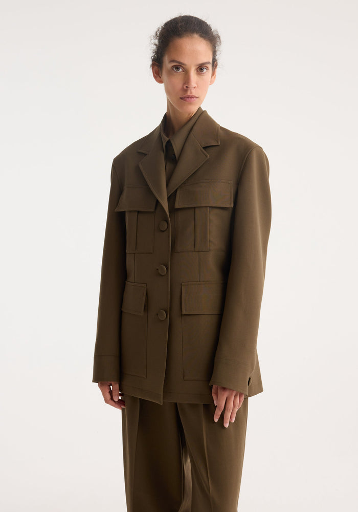 Model wearing Safari blazer-jacket | forest, featuring a boxy fit, notched collar, four flap pockets, and cloth-covered buttons, paired with loose-cut trousers.