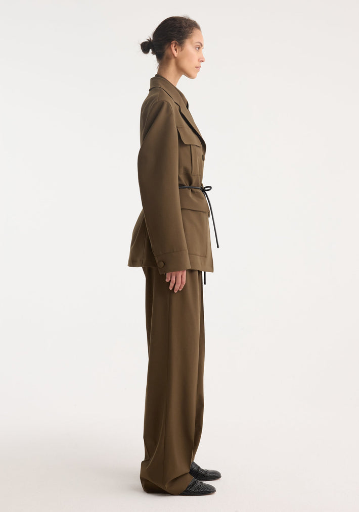Model in side profile wearing a Safari blazer-jacket | forest, featuring a belted design, four flap pockets, and wide-leg trousers, styled with slip-on shoes.