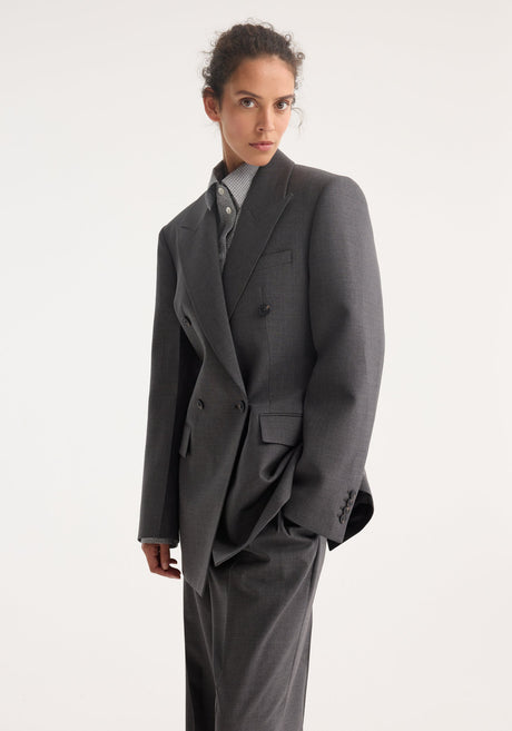 Model wearing the Hourglass blazer in dark grey melange, featuring a sharp notch lapel, strong shoulders, cinched waist, over matching wide-leg trousers.