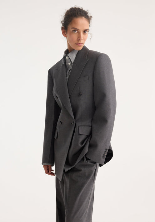 Model wearing the Hourglass blazer in dark grey melange, featuring a sharp notch lapel, strong shoulders, cinched waist, over matching wide-leg trousers.