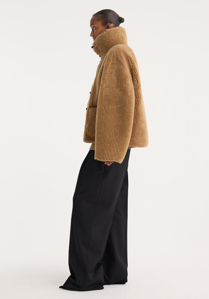 Profile view of a person in a Shearling coat | cognac, featuring a stand-up collar, wide-leg trousers, leather mandarin closures, and patch pockets.