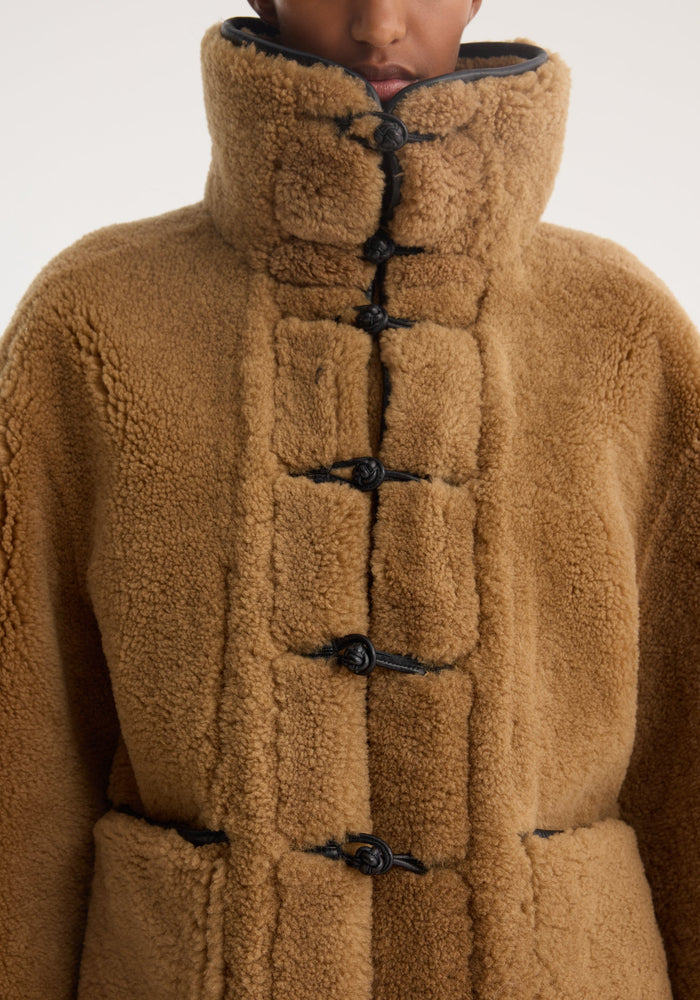 Shearling coat | cognac: Close-up of a plush, camel-colored shearling coat with black toggle closures, high collar, and leather trim, worn by a model.
