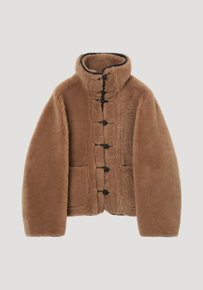 Shearling coat | cognac features a plush, camel brown texture, mandarin closures, stand-up collar, patch pockets, and leather trim, offering a cozy, oversized style.