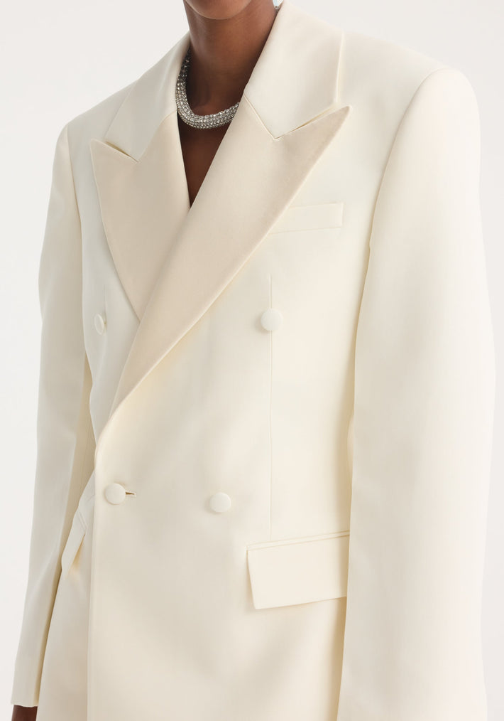 Woman wearing Hourglass tuxedo blazer in off white, featuring peaked lapels, satin details, covered buttons, and flap pockets, paired with a sparkling choker.