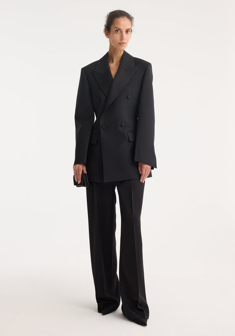 Woman wearing Hourglass tuxedo blazer with sharp notch lapels, strong shoulders, and cinched waist, paired with matching high-waisted trousers, holding a black clutch.