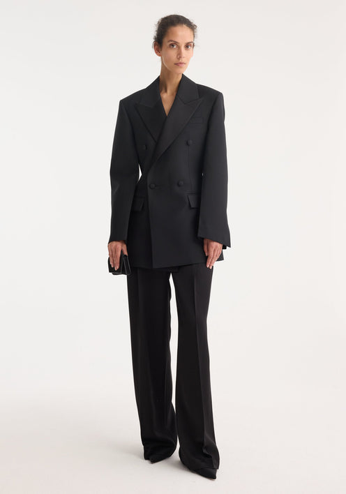 Hourglass tuxedo blazer in black with peaked satin lapels, flap pockets, and covered buttons, showcasing a sculptural silhouette with a cinched waist and strong shoulders.