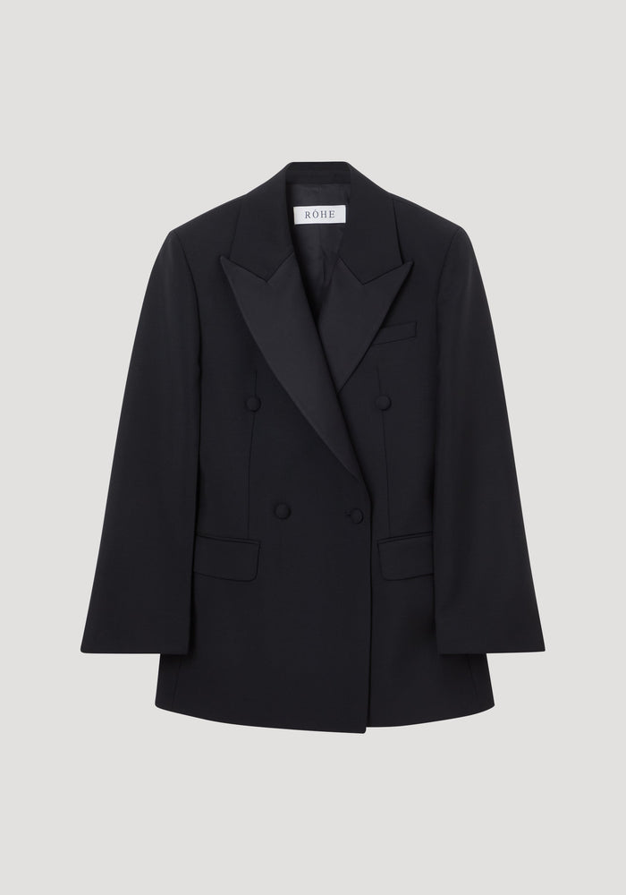 Hourglass tuxedo blazer in black with peaked satin lapels, flap pockets, and covered buttons, showcasing a sculptural silhouette with a cinched waist and strong shoulders.