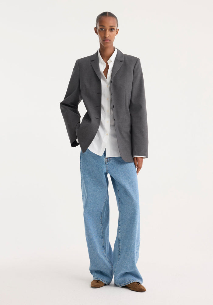Woman in fitted wool blazer, dark grey melange, styled with white shirt, wide-leg jeans, and loafers; hands in pocket, showcasing modern tailored look.