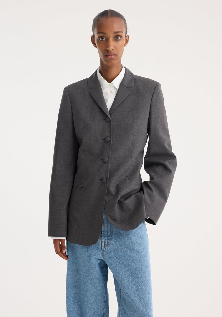 Model wearing a fitted wool blazer in dark grey melange with fabric-covered buttons and welt pockets, paired with loose-fit light-wash jeans, hair pulled back.