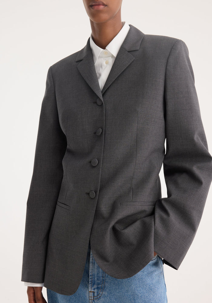 Person wearing a fitted wool blazer in dark grey melange with covered buttons, notch lapel, and welt pockets, paired with a white shirt and blue jeans.
