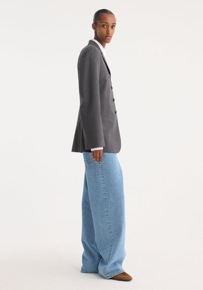 Person in profile wearing a fitted wool blazer in dark grey melange, featuring notch lapels, covered buttons, and welt pockets, styled over a white shirt.