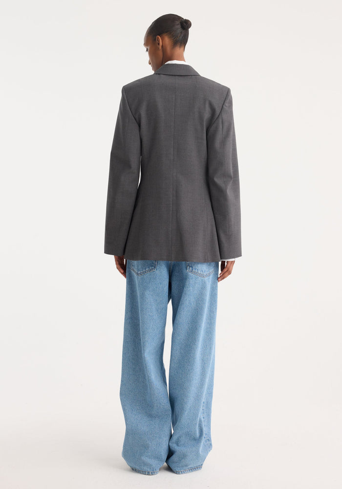 Person wearing a fitted wool blazer in dark grey melange, featuring notch lapels and covered buttons, paired with loose-fitting light blue jeans, hair in a bun.