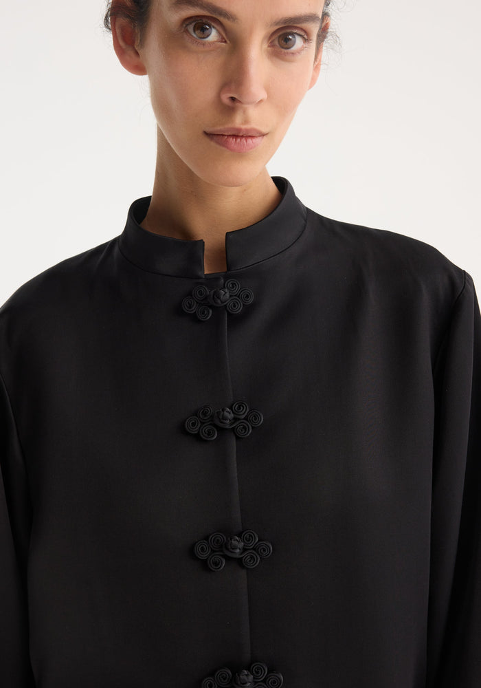 Close-up of a woman wearing a black Mandarin satin jacket with a mandarin collar and pankou closures. The jacket is made of 100% viscose.