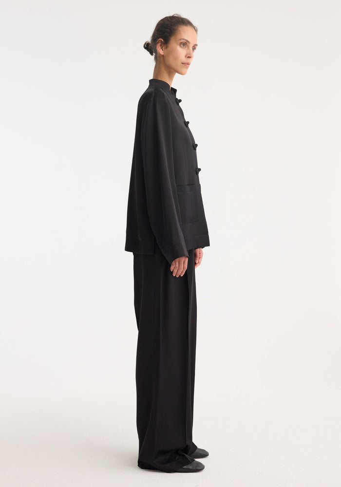Model in side profile wearing a Mandarin satin jacket | black, featuring pankou closures and patch pockets, paired with matching wide-leg trousers and flat shoes.