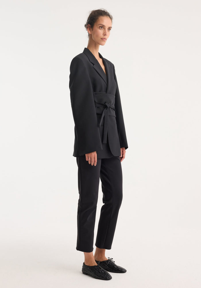Woman models a Kimono belted blazer in black, featuring a wrap sash belt and tailored fit, paired with slim trousers and lace-up loafers.